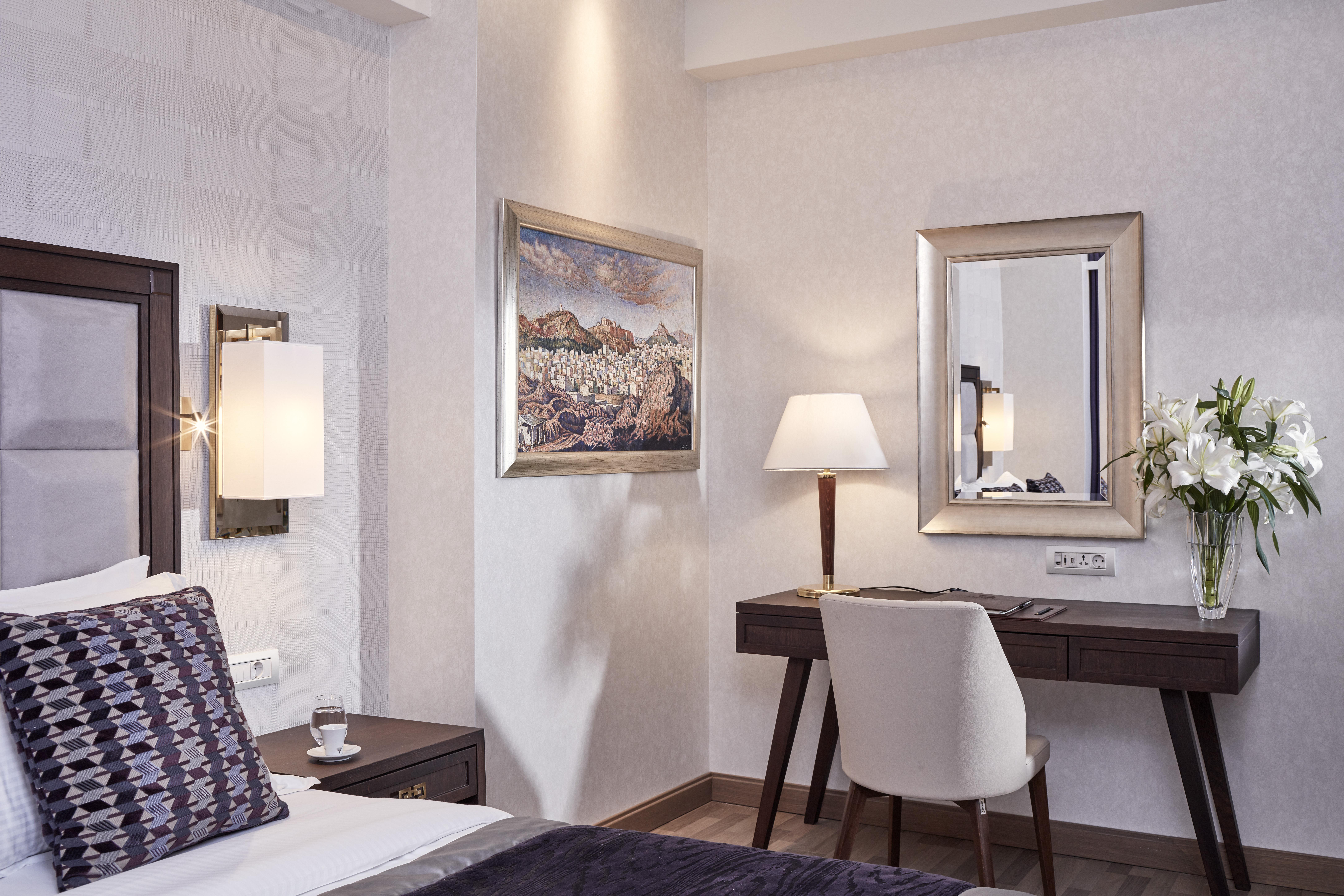 Electra Metropolis Hotell Athen Eksteriør bilde The photo shows a well-decorated hotel room. On the left, there is a bed with patterned pillows, and on the right, a desk set against the wall. The desk has a modern chair and is accompanied by a lamp and a mirror. Above the desk, there's a piece of 