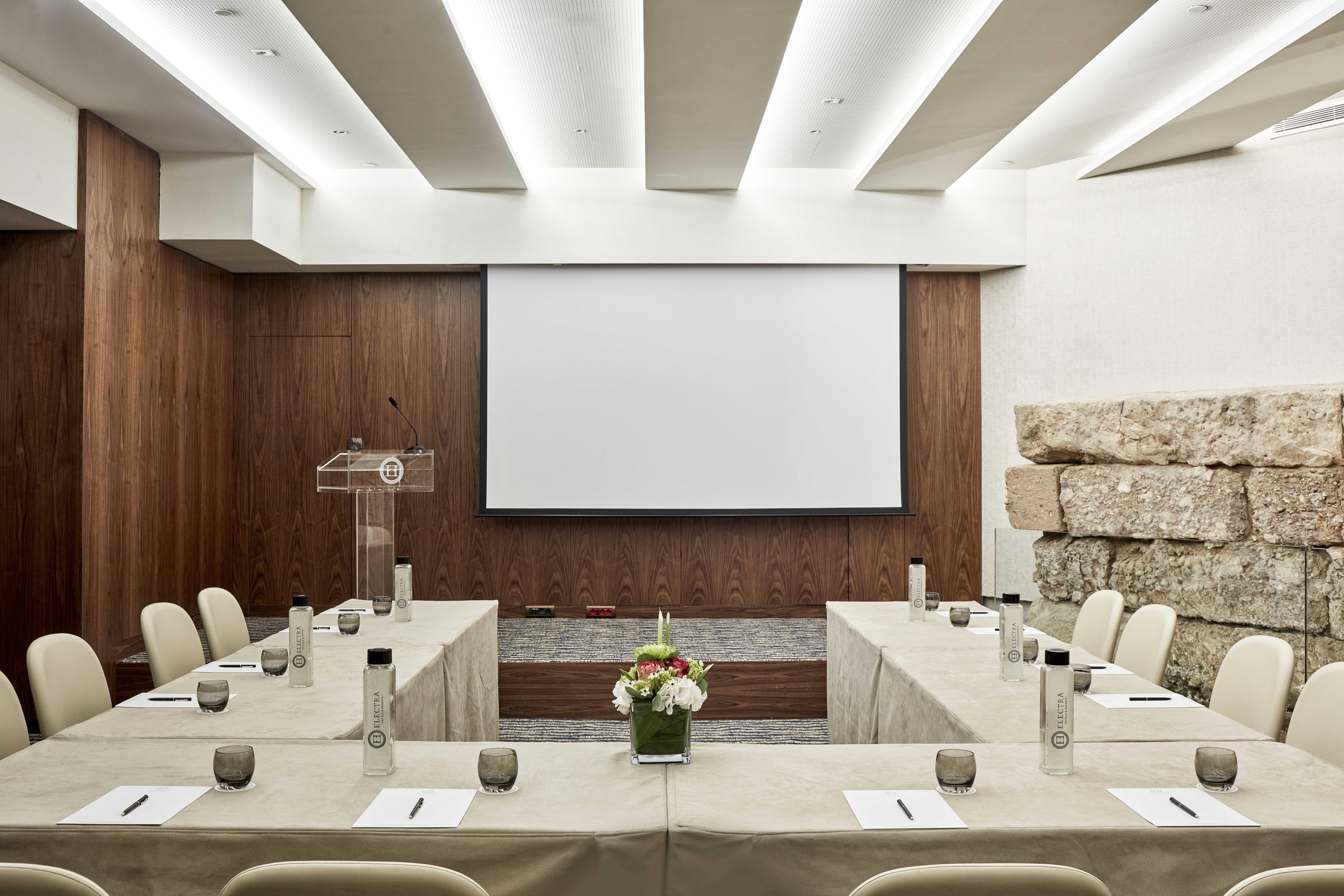 Electra Metropolis Hotell Athen Eksteriør bilde The image shows a modern conference room setup. The room features a large screen at the front, likely for presentations. There are several tables arranged in a U-shape, covered with light-colored tablecloths, and each table has glasses and notepads. 