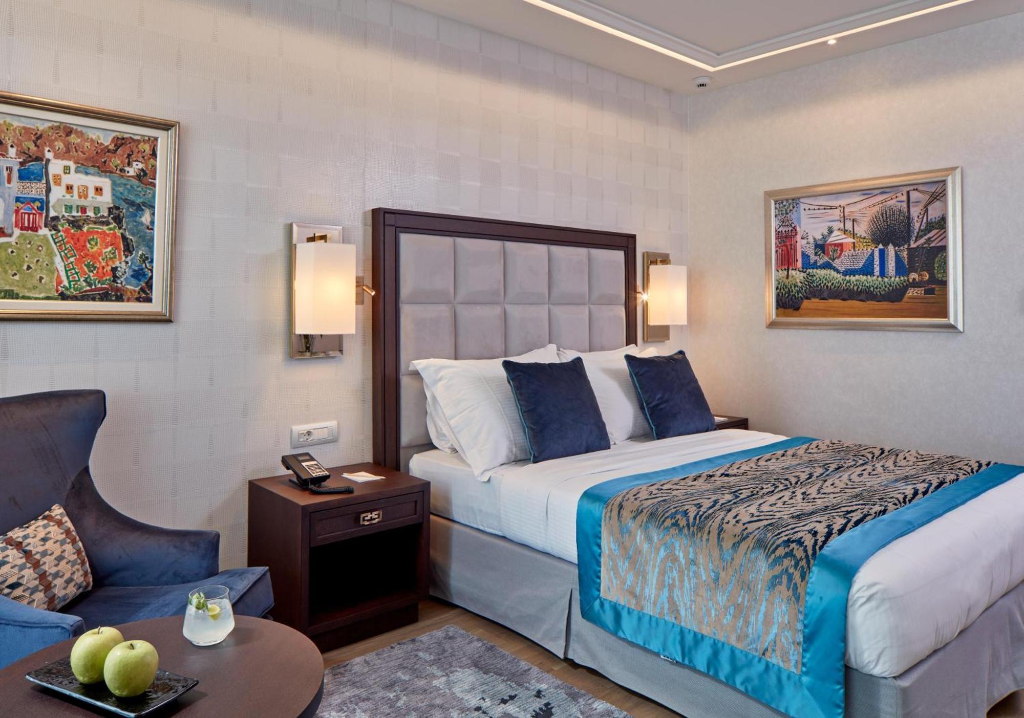 Electra Metropolis Hotell Athen Eksteriør bilde The photo shows a modern hotel room featuring a double bed with decorative pillows. The bed has a stylish headboard, and the bedding includes a patterned throw. Beside the bed, there are bedside lamps and a wooden nightstand with a phone. 

On the le