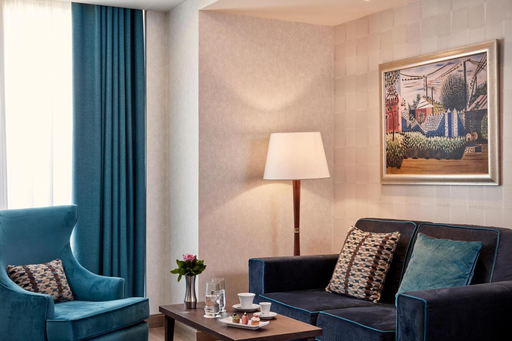 Electra Metropolis Hotell Athen Eksteriør bilde The photo shows a stylish and modern living space. There is a dark blue couch adorned with decorative cushions, paired with a comfortable blue armchair. A coffee table in the center features a small floral arrangement and a couple of glassware items,