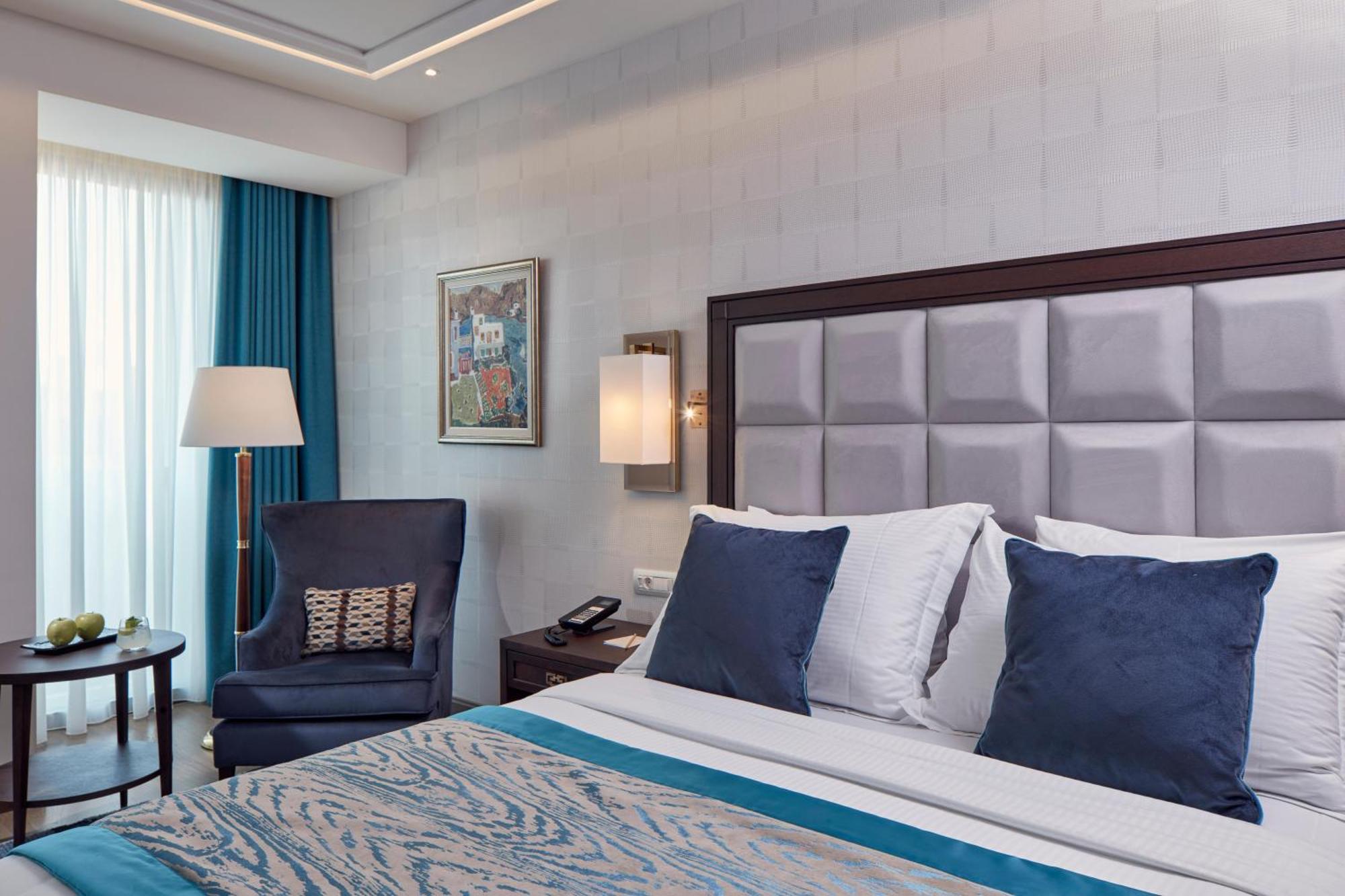 Electra Metropolis Hotell Athen Eksteriør bilde The photo depicts a modern hotel room featuring a neatly made bed with white linens and blue accent pillows. There's a plush blue armchair next to a small table, which may hold some decorative items, including a bowl of green apples. A large window a