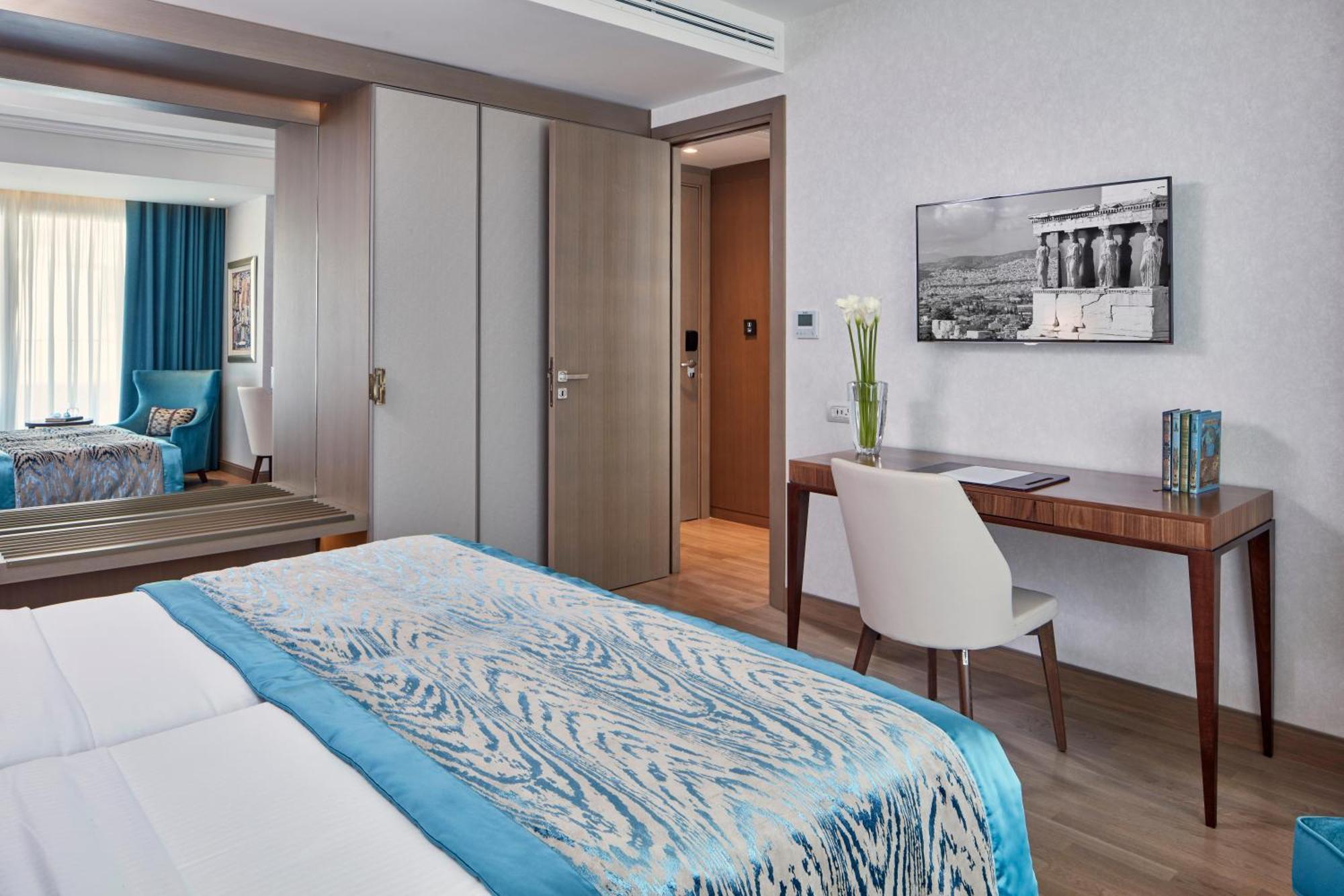Electra Metropolis Hotell Athen Eksteriør bilde The photo shows a modern hotel room with two beds covered in light-colored linens that have a blue patterned throw or blanket. There's a wooden desk and a white chair situated next to the wall, with a vase of flowers on the desk and a framed black-an