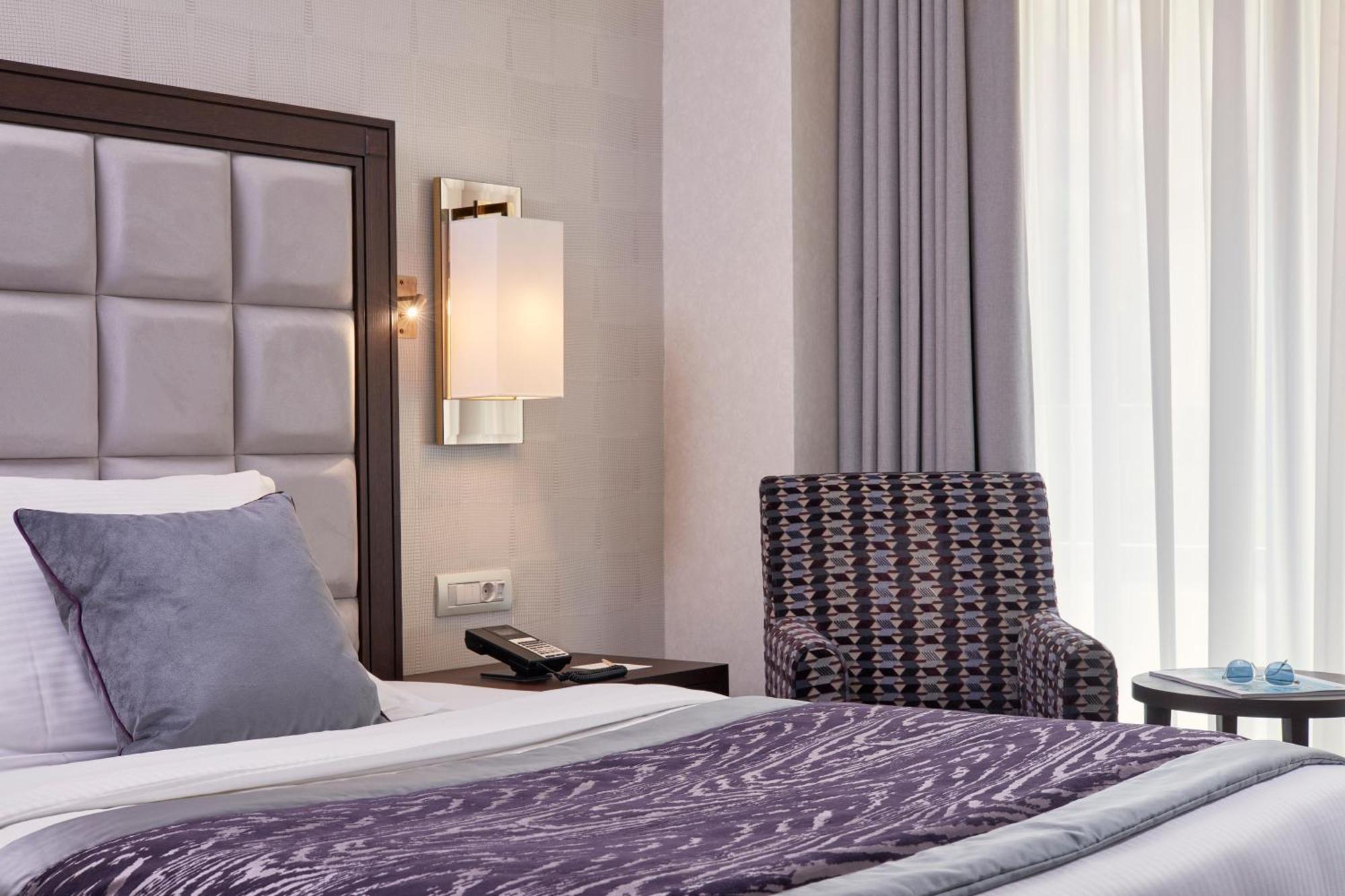 Electra Metropolis Hotell Athen Eksteriør bilde The photo shows a modern hotel room featuring a neatly made bed with a dark purple throw blanket and large pillows. Behind the bed, there is a stylish headboard with a tufted design. A soft, ambient light fixture is mounted on the wall beside the bed