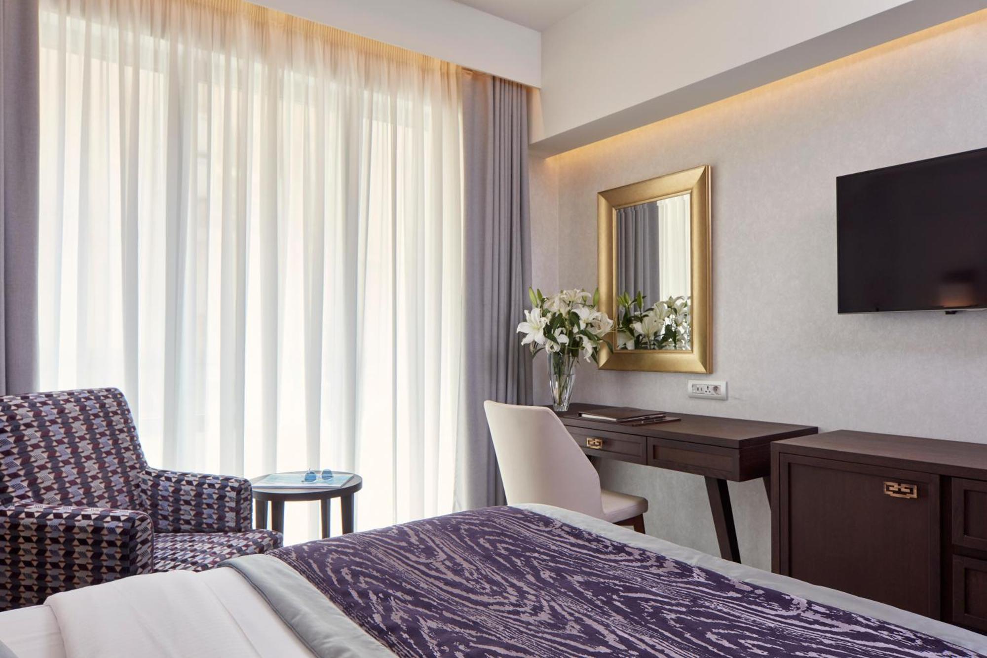 Electra Metropolis Hotell Athen Eksteriør bilde The photo shows a well-furnished hotel room. On the left, there is a patterned chair with a small round table next to it. The room features large windows covered with sheer curtains that allow natural light to fill the space. In the center, there is 