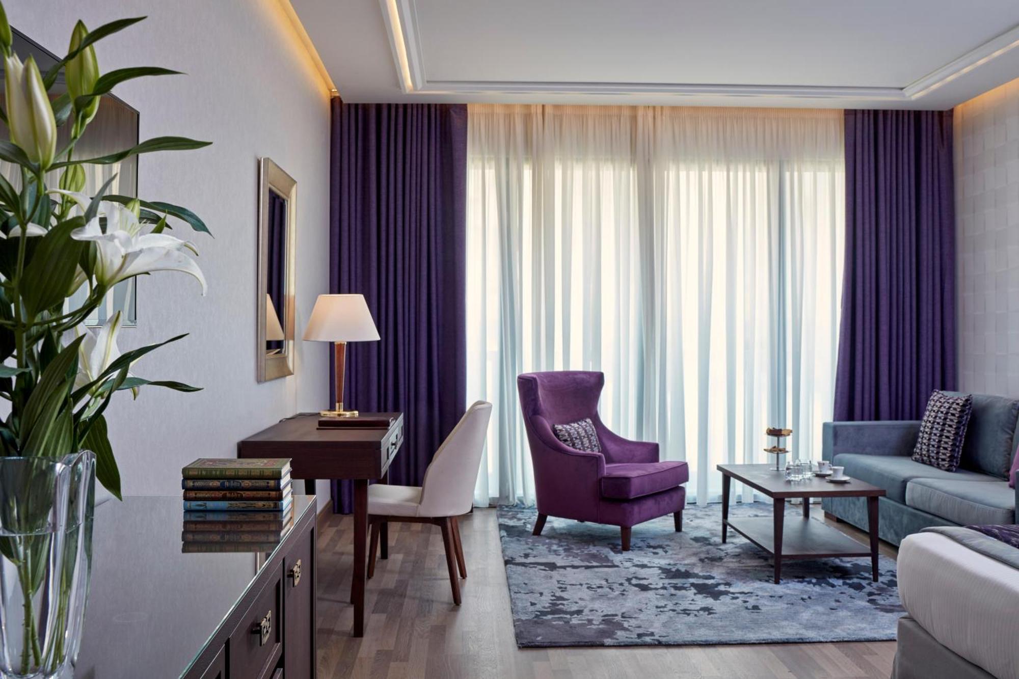 Electra Metropolis Hotell Athen Eksteriør bilde The photo depicts a stylish and modern living space. On one side, there's a plush purple armchair and a small wooden side table with a lamp. Nearby, a light-colored sofa is placed with a coffee table in front of it. The room features large windows co