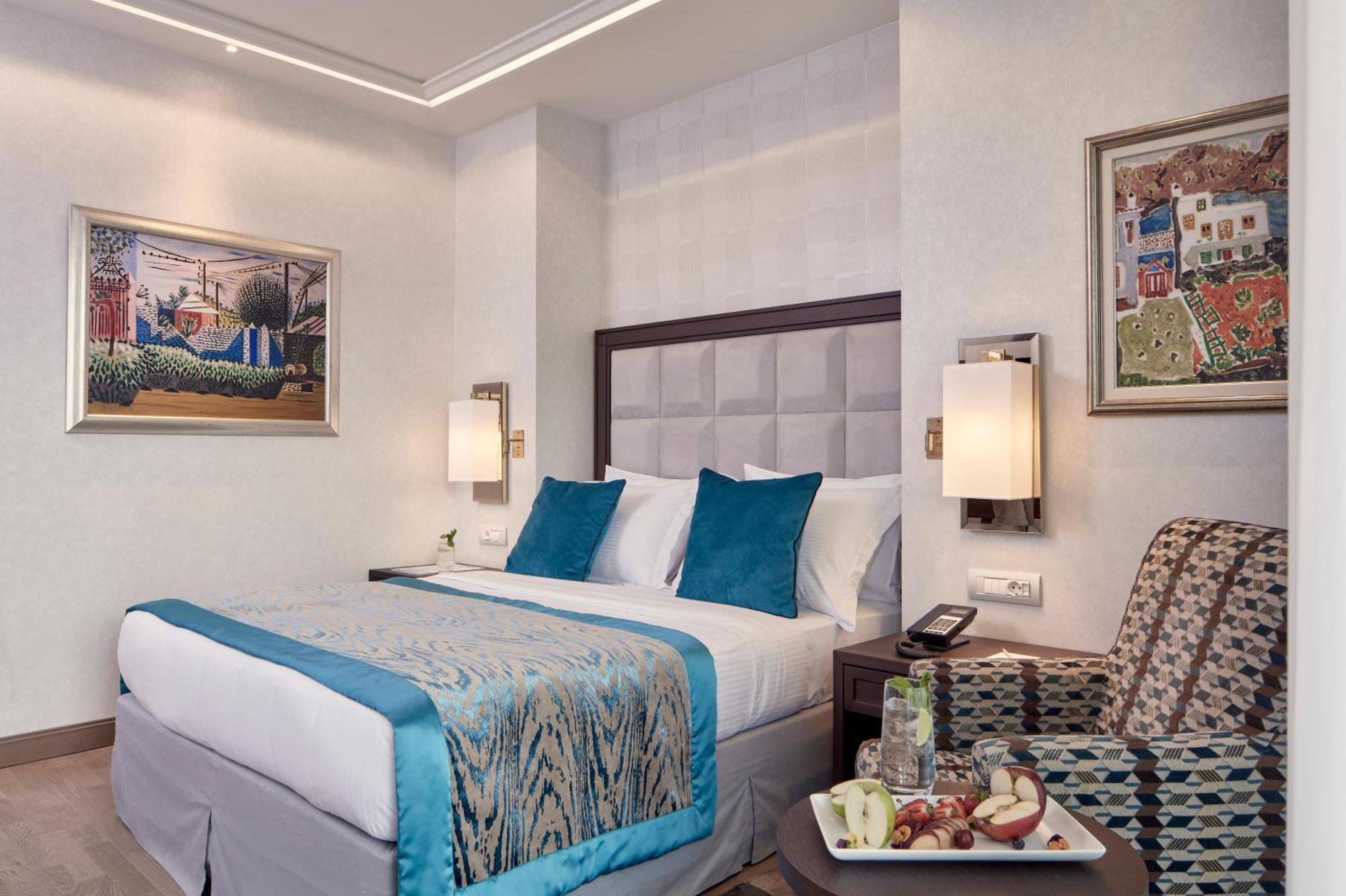 Electra Metropolis Hotell Athen Eksteriør bilde The photo shows a hotel room featuring a queen-sized bed with a light blue and patterned bedspread. The bed has white pillows with two accent pillows in turquoise. Beside the bed, there are contemporary lamps mounted on the wall. 

To the side, there