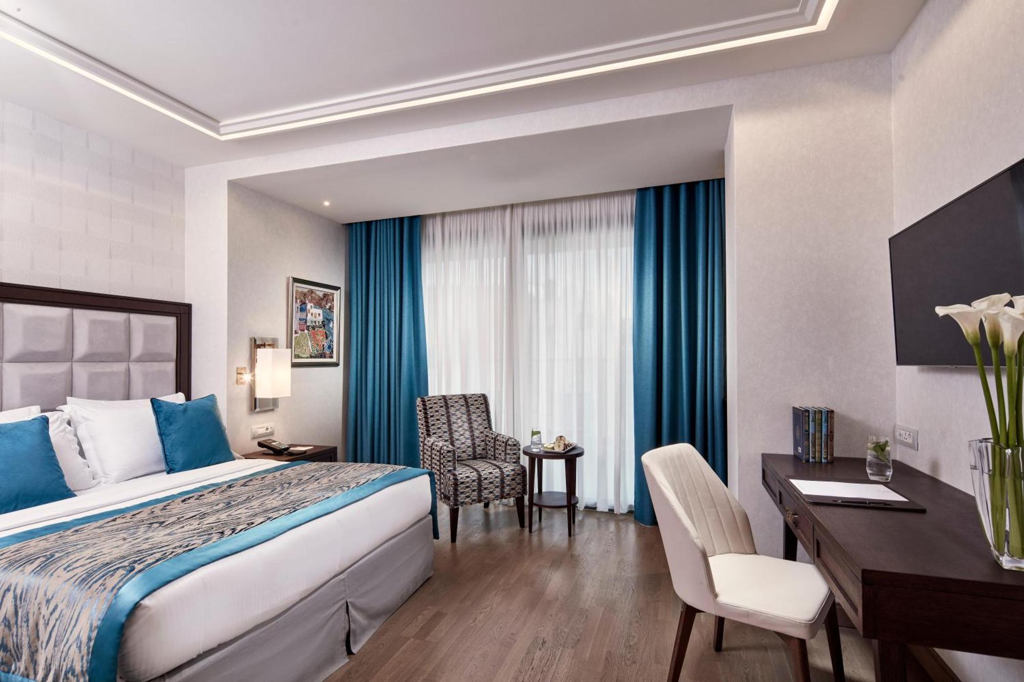 Electra Metropolis Hotell Athen Eksteriør bilde The photo shows a well-decorated hotel room. It features a large bed with a blue and neutral colored bedding set. There are two blue curtains that frame a window, allowing natural light to enter the space. Next to the bed, there's a small chair and t