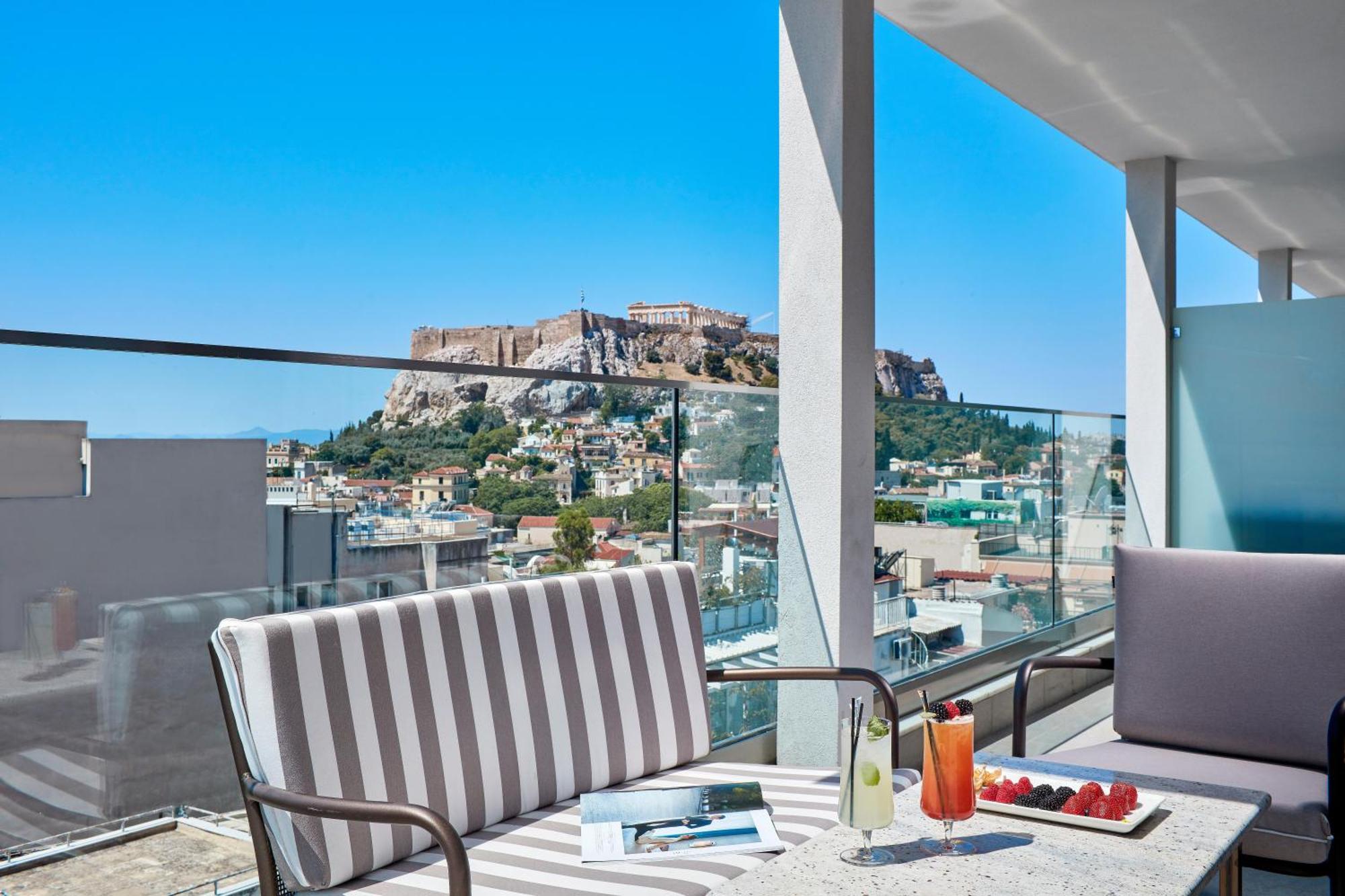 Electra Metropolis Hotell Athen Eksteriør bilde The photo features a modern terrace or balcony with stylish seating arrangements. There are two cushioned chairs, one with striped fabric. A table in front holds a beverage, possibly a cocktail, and some fresh strawberries. The backdrop showcases a b
