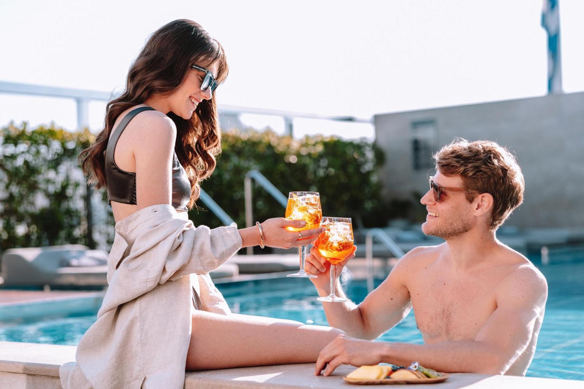 Electra Metropolis Hotell Athen Eksteriør bilde The photo shows a pair enjoying a vibrant moment by a pool. A woman, sitting on the edge, is wearing stylish sunglasses and a bikini, and she is handing a drink to a man sitting beside her. He is shirtless, also wearing sunglasses, and is smiling whi